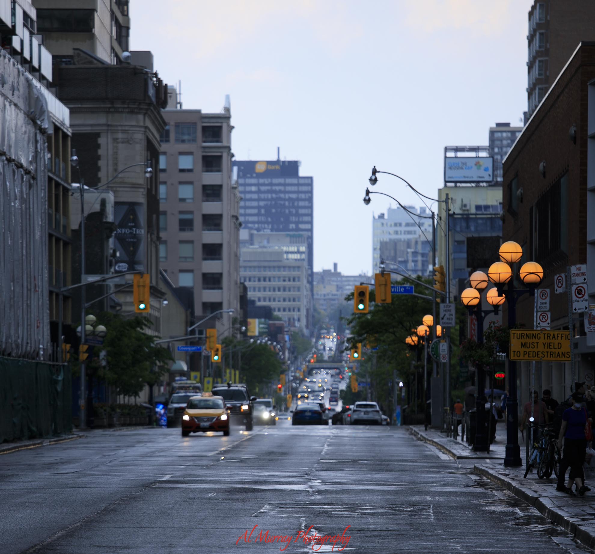 Rain falls on a busy downtown Toronto street with wet pavement. Cars move between tall buildings while glowing street lamps line the sidewalk.