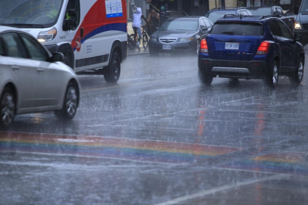things to do in Toronto indoors Heavy rain falls on a busy Toronto street as cars drive through water covered pavement beside a rainbow painted crosswalk. The wet city scene sets the context for finding things to do in Toronto on a rainy day.