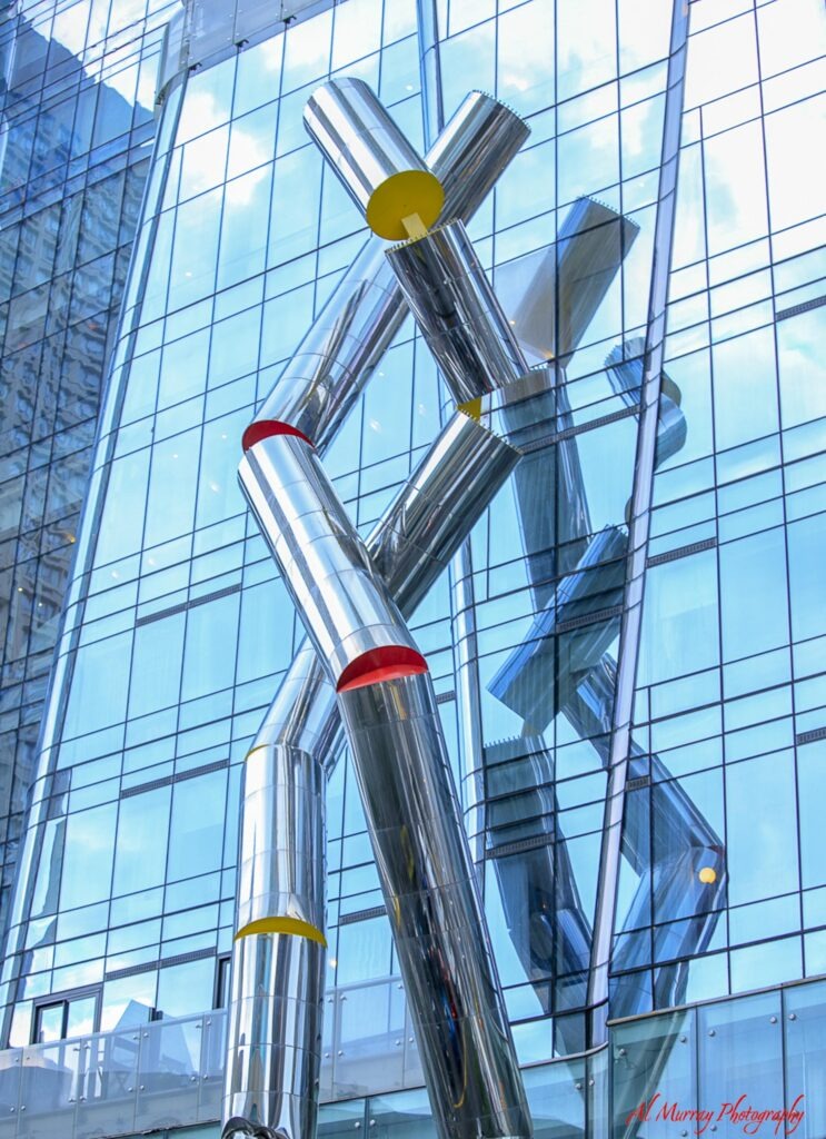 Large abstract sculpture made of shiny silver metal tubes with red and yellow accents stands in front of a modern glass building reflecting the blue sky. The striking contemporary artwork adds to the upscale atmosphere of the Bloor Yorkville shopping district in Toronto.